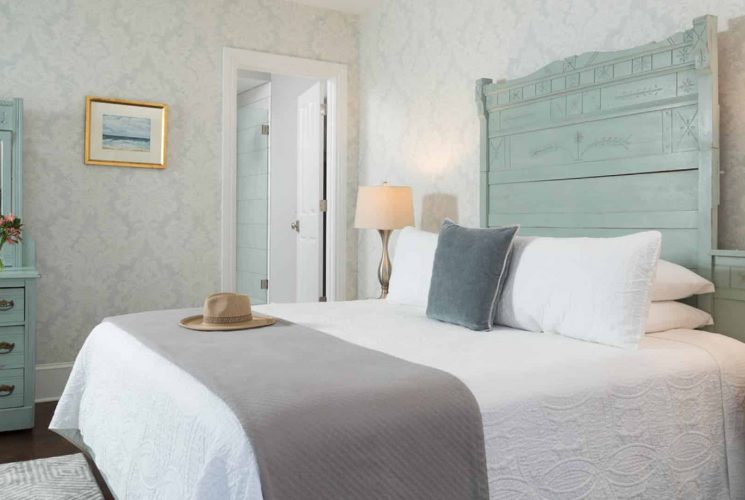 Bedroom with large ornate seafoam headboard, matching dresser with mirror, white bedding, gray blanket, hardwood flooring, and light brocade wallpaper