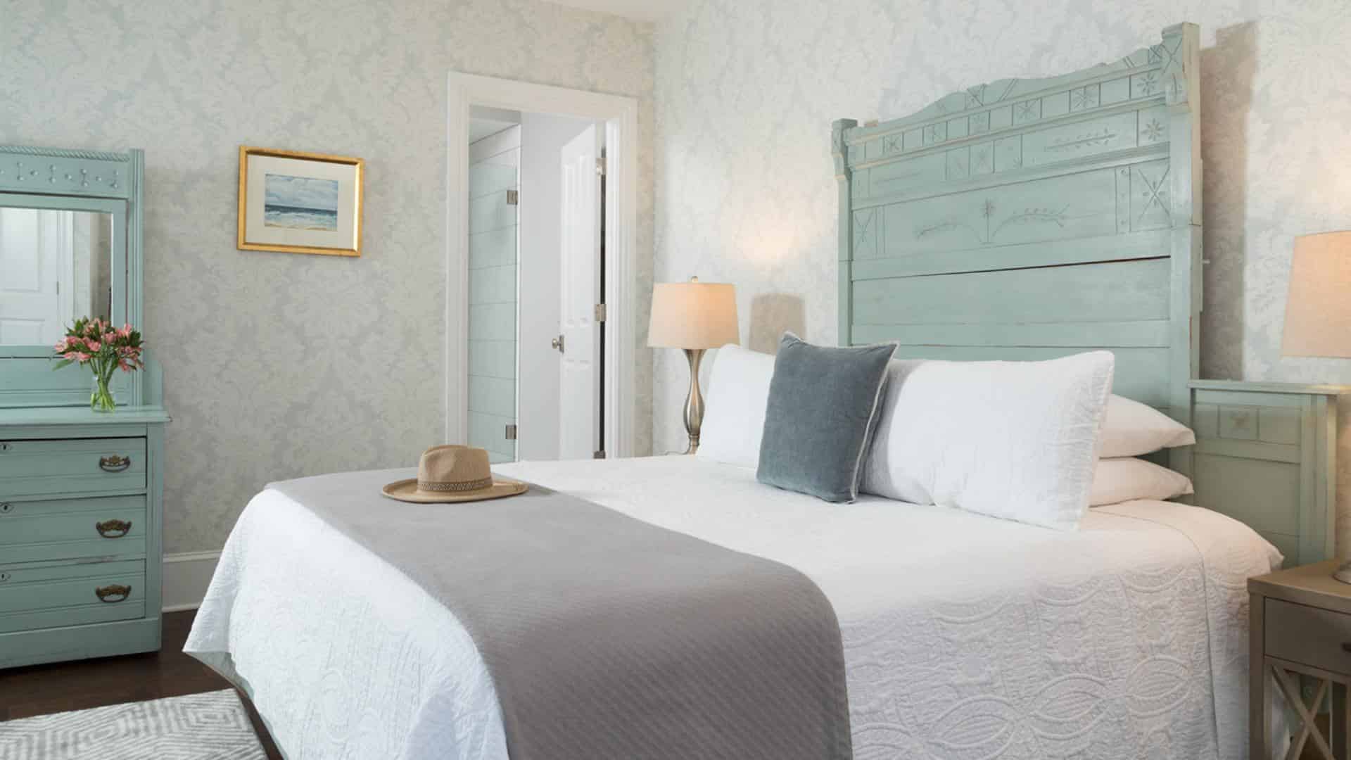 Bedroom with large ornate seafoam headboard, matching dresser with mirror, white bedding, gray blanket, hardwood flooring, and light brocade wallpaper