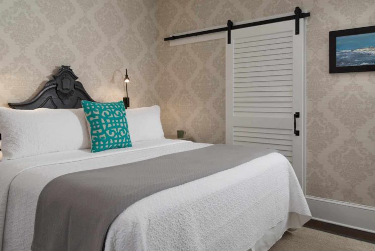 Bedroom with dark wooden ornate headboard, white bedding, gray blanket, hardwood flooring, and ornate wallpaper