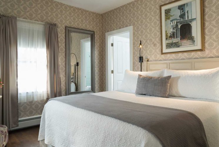 Bedroom with light-colored wooden headboard, white bedding, gray blanket, hardwood flooring, sitting area with upholstered chair, and large mirror