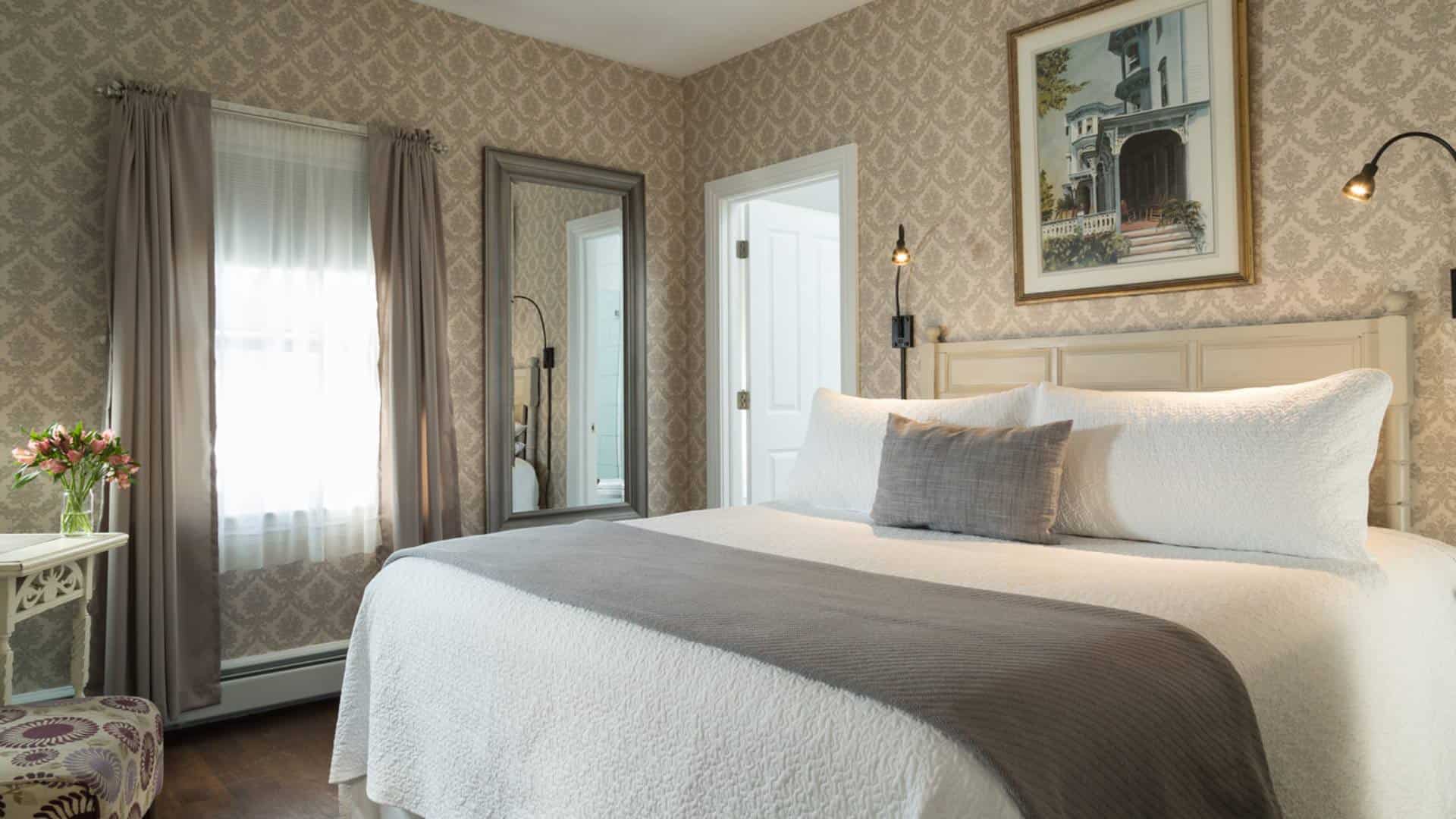 Bedroom with light-colored wooden headboard, white bedding, gray blanket, hardwood flooring, sitting area with upholstered chair, and large mirror