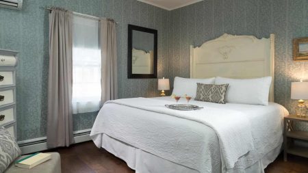 Bedroom with large light-colored ornate headboard, white bedding, hardwood flooring, and sitting area with upholstered chair