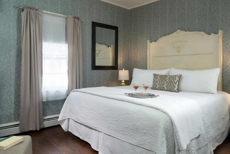 Bedroom with large light-colored ornate headboard, white bedding, hardwood flooring, and sitting area with upholstered chair