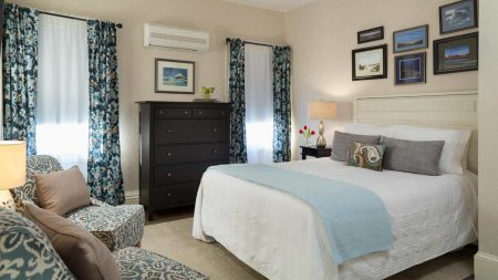 Large bedroom with light-colored ornate headboard, white bedding, blue blanket, dark wooden dresser, and sitting area with upholstered chairs