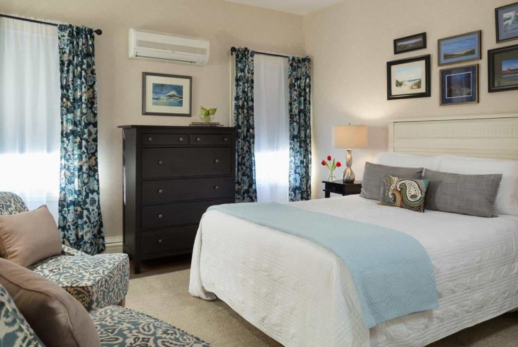 Large bedroom with light-colored ornate headboard, white bedding, blue blanket, dark wooden dresser, and sitting area with upholstered chairs