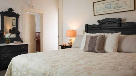 Bedroom with dark wooden ornate headboard, matching dresser with mirror, cream-colored bedding, and dark wooden nightstand