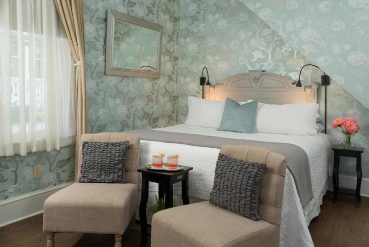 Bedroom with antiqued headboard, white bedding, sitting area with tan upholstered chairs, hardwood flooring, and ornate wallpaper