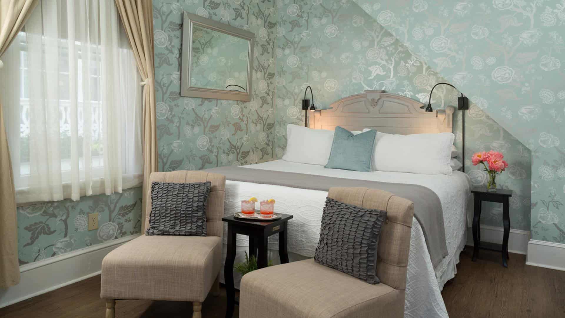 Bedroom with antiqued headboard, white bedding, sitting area with tan upholstered chairs, hardwood flooring, and ornate wallpaper