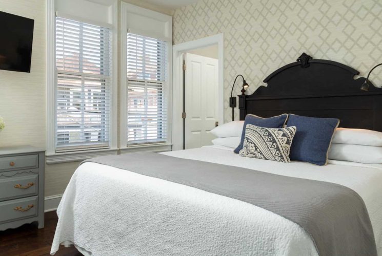 Bedroom with dark wooden headboard, white bedding, gray blanket, hardwood flooring, gray dresser, and modern wallpaper