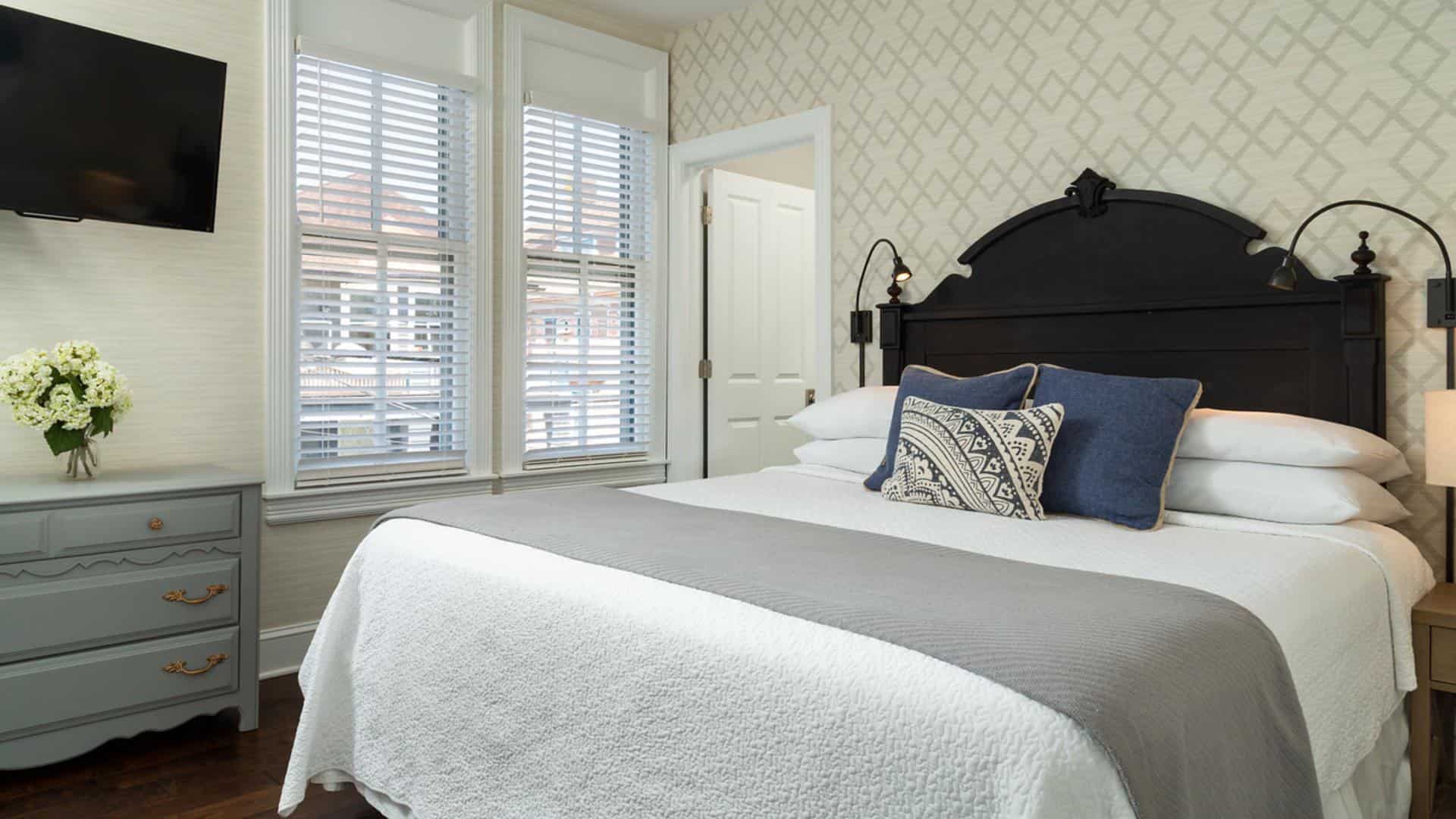 Bedroom with dark wooden headboard, white bedding, gray blanket, hardwood flooring, gray dresser, and modern wallpaper