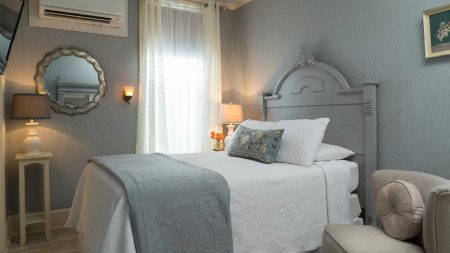 Bedroom with gray wooden headboard, white bedding, hardwood flooring, light gray patterned wallpaper, small upholstered chair, and mirror on the wall