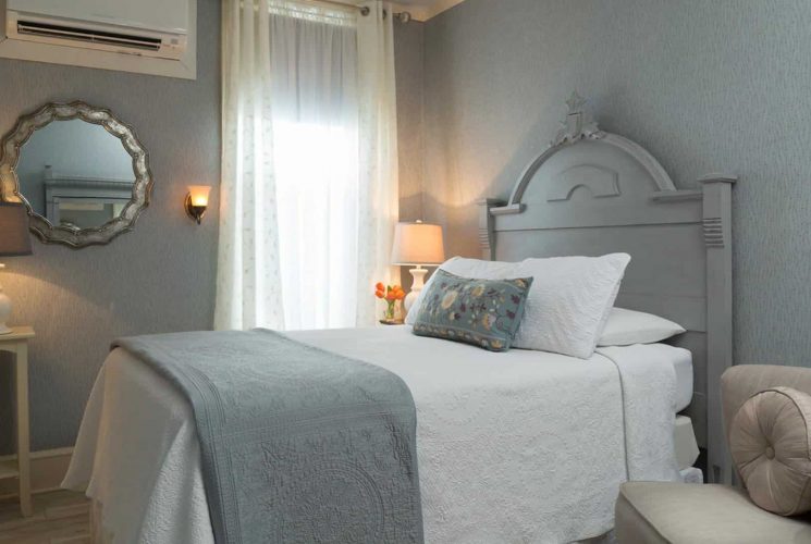 Bedroom with gray wooden headboard, white bedding, hardwood flooring, light gray patterned wallpaper, small upholstered chair, and mirror on the wall