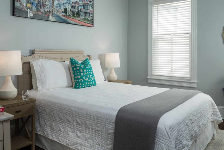 Bedroom with light-colored ornate headboard, white bedding, gray blanket, hardwood flooring, and light gray walls