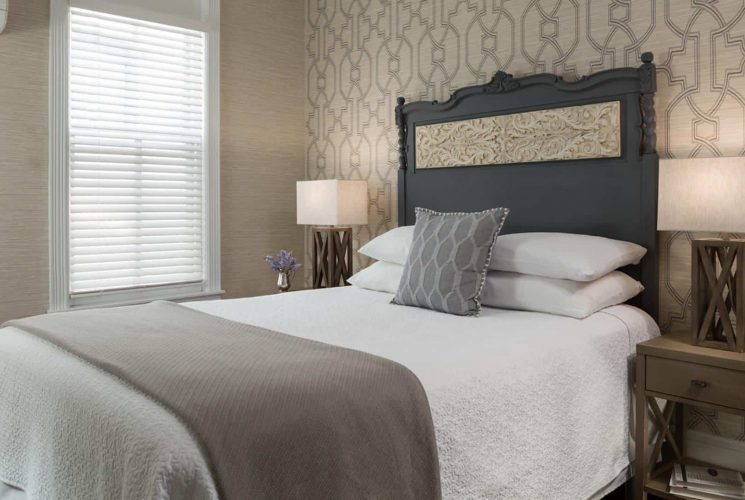 Bedroom with large dark wooden ornate headboard, white bedding, gray blanket, light-tan and modern wallpaper, and wooden nightstands