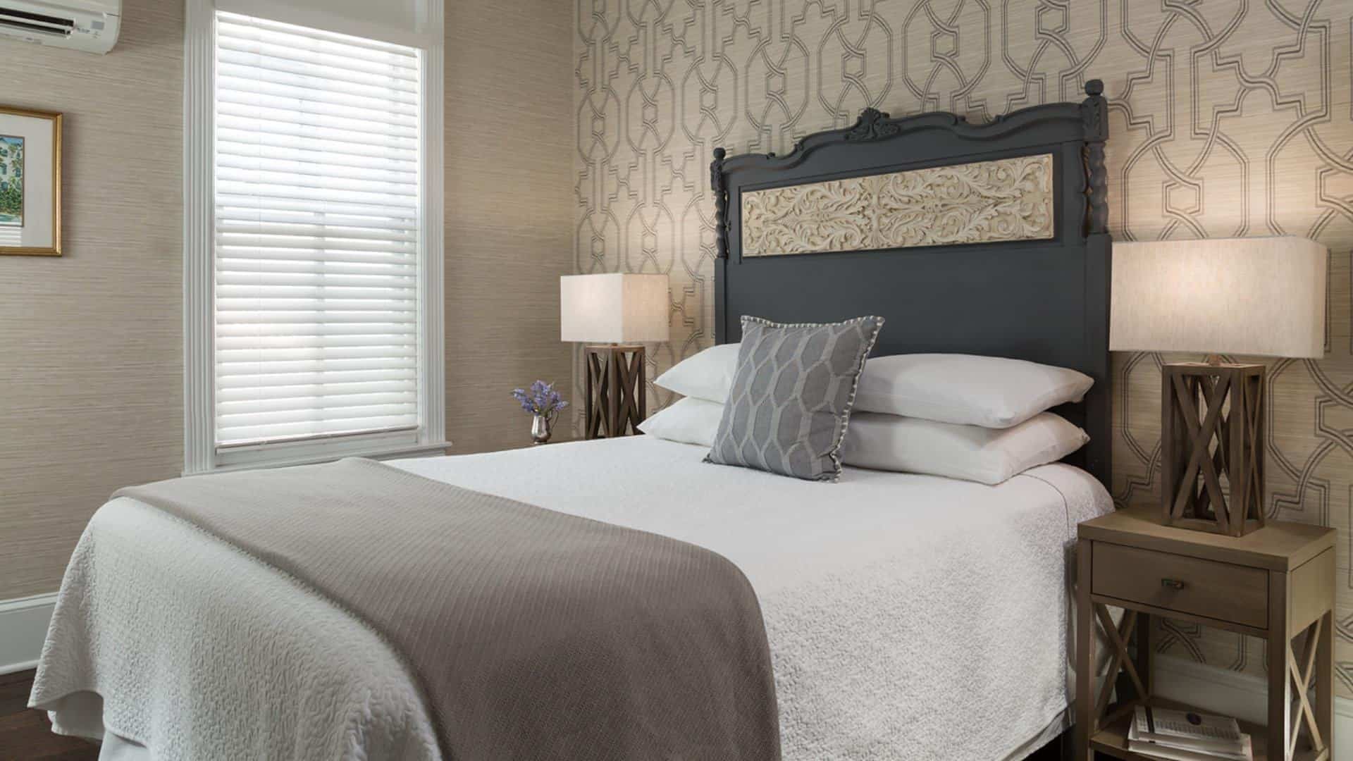 Bedroom with large dark wooden ornate headboard, white bedding, gray blanket, light-tan and modern wallpaper, and wooden nightstands