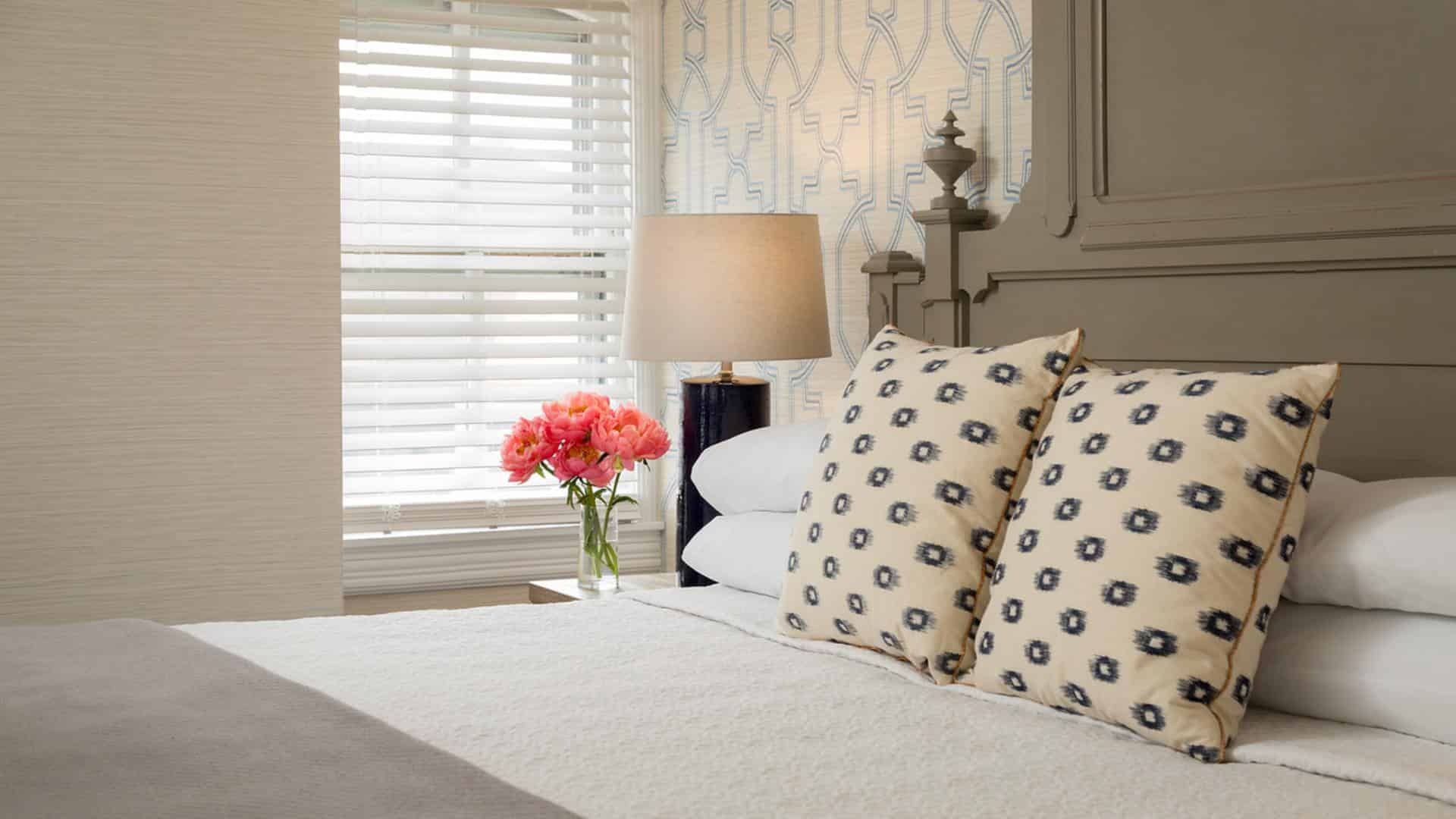 Bedroom with large gray headboard, white bedding, gray blanket, yellow and black pillows, and vase full of pink flowers