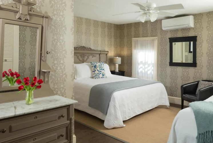 Large bedroom with gray wooden ornate headboard, matching dresser with marble top and mirror, white bedding, hardwood flooring, and brocade wallpaper