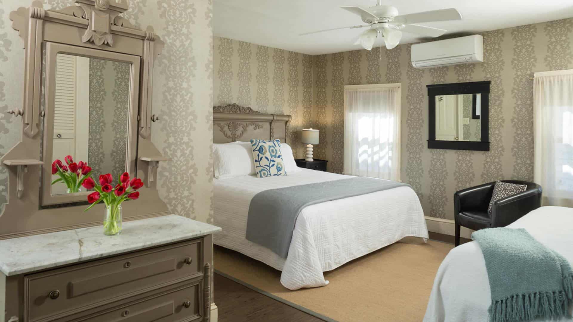 Large bedroom with gray wooden ornate headboard, matching dresser with marble top and mirror, white bedding, hardwood flooring, and brocade wallpaper