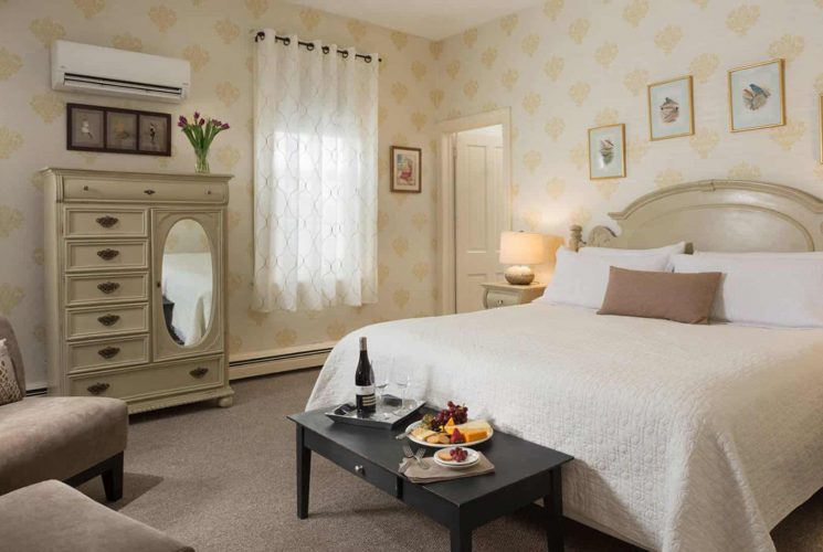 Large bedroom with antiqued headboard, white bedding, sitting area with tan upholstered chairs, and antiqued wooden dresser