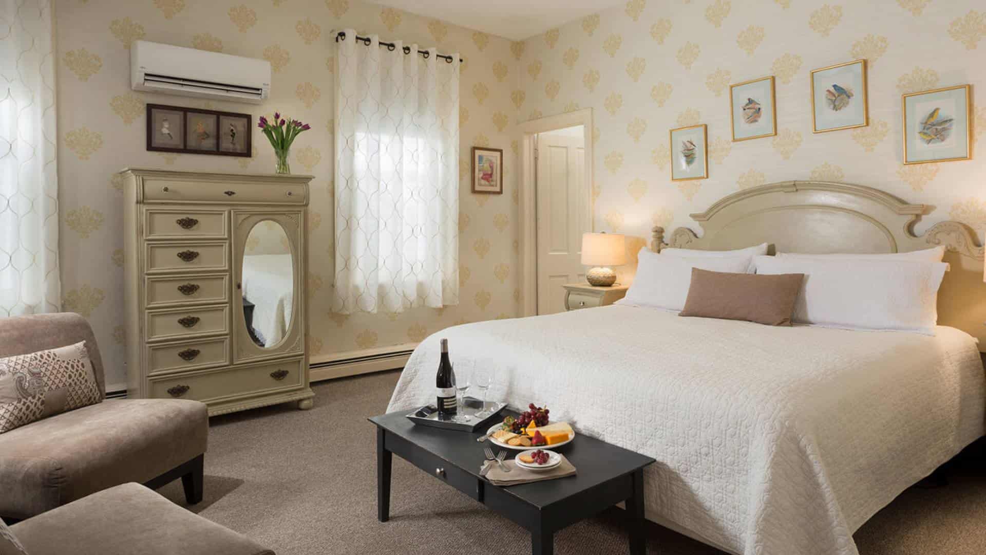 Large bedroom with antiqued headboard, white bedding, sitting area with tan upholstered chairs, and antiqued wooden dresser