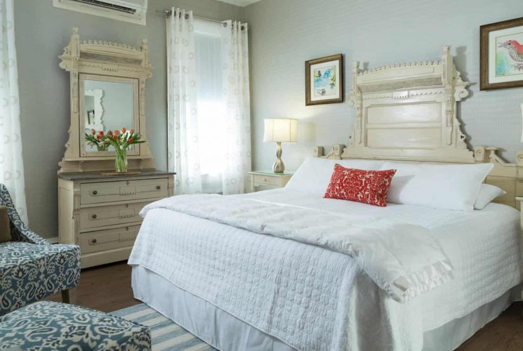 Large bedroom with ornate antique headboard, white bedding, hardwood flooring, sitting area with upholstered chairs, and antique dresser with mirror