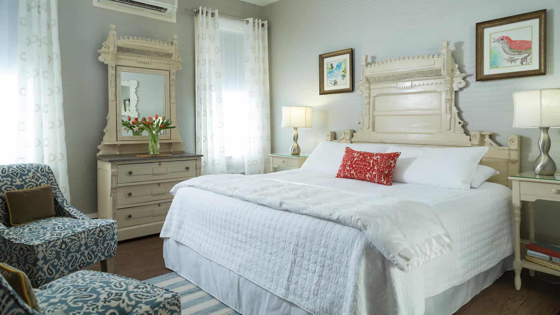 Large bedroom with ornate antique headboard, white bedding, hardwood flooring, sitting area with upholstered chairs, and antique dresser with mirror