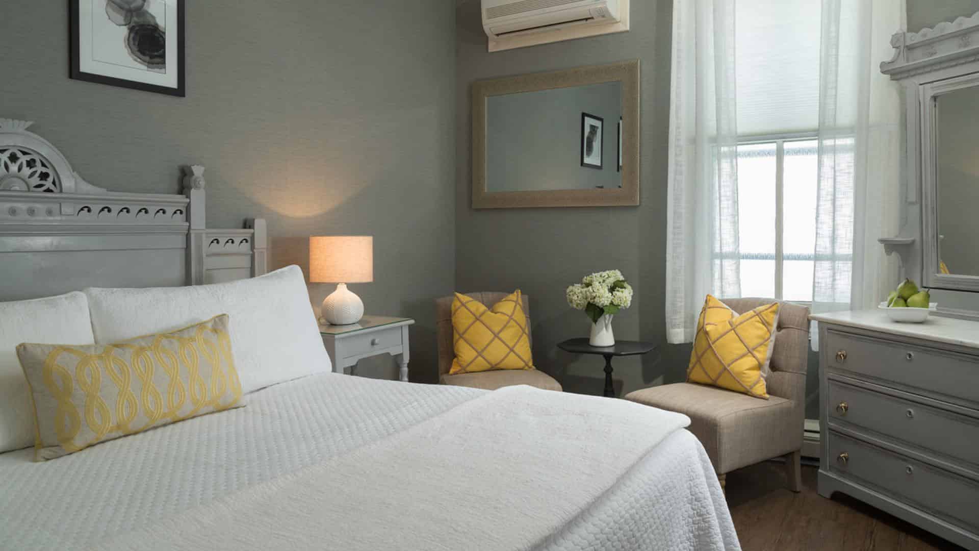 Bedroom with large gray headboard, white bedding, yellow pillows, hardwood flooring, gray walls, and gray dresser with marble top and mirror