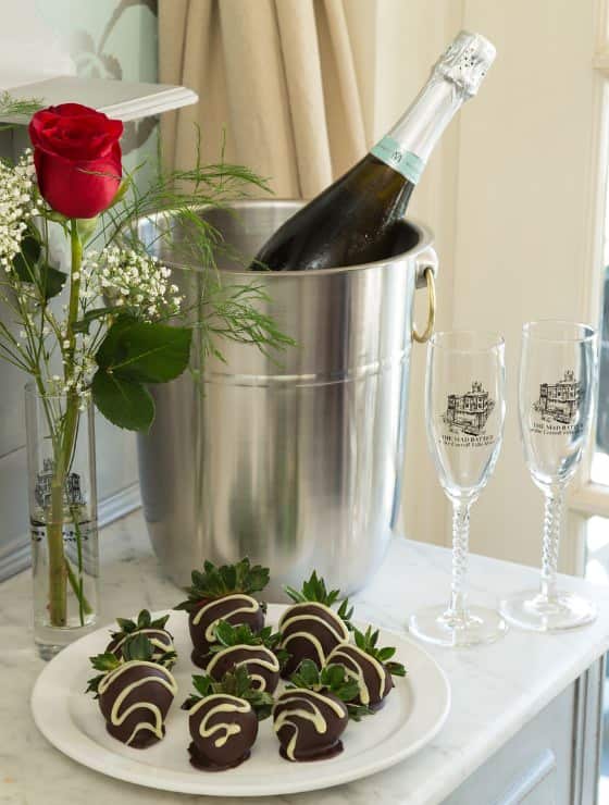 White porcelain plate with chocolate covered strawberries, vase with single red rose, two empty champagne flutes, silver ice bucket with chilled Champagne