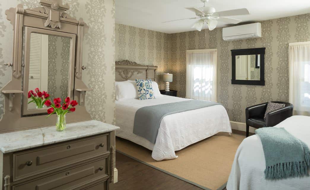 Large bedroom with dark wooden hardwood flooring, brocade wallpaper, white bedding, taupe dresser with marble top, and brown leather armchair