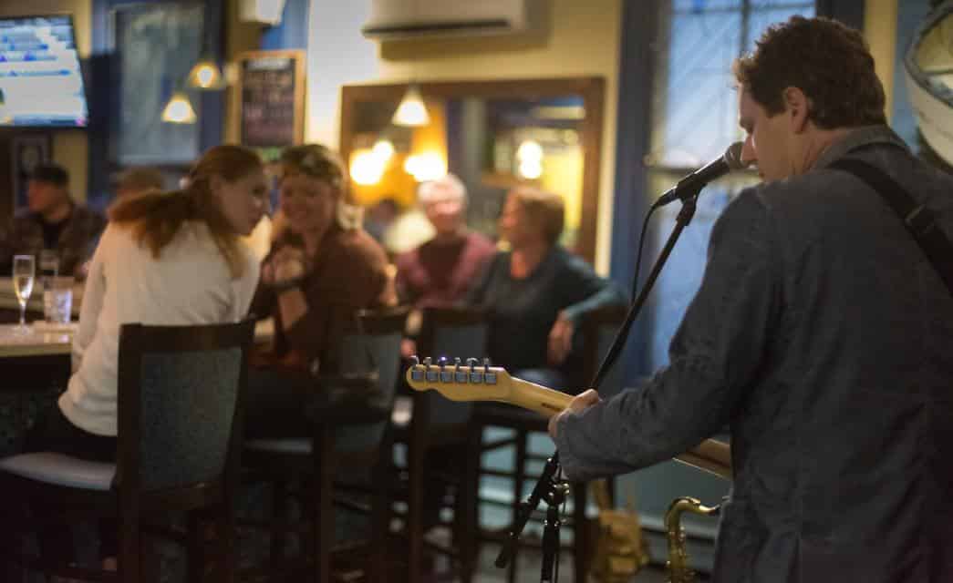 Man playing a guitar and singing in a bar with people sitting and watching in the background