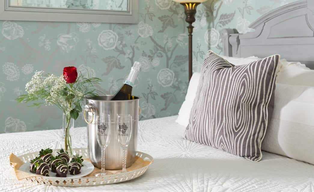 Close up view of white plate with chocolate covered strawberries and glasses filled with wine sitting on tray on bed with white bedding