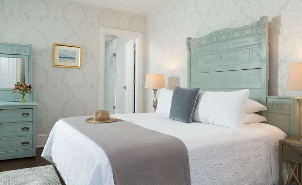 Bedroom with ornate antique headboard, white bedding, hardwood flooring, and antique dresser with mirror