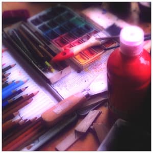 A table full of art supplies - red paint, colored pencils, paper, paints and paintbrushes