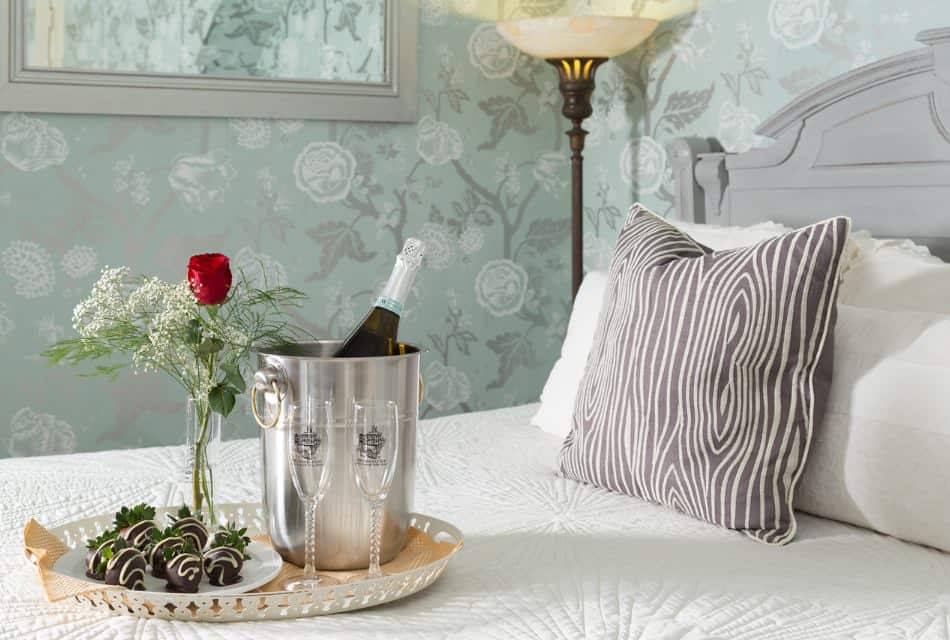 Close up view of white plate with chocolate covered strawberries and glasses filled with wine sitting on tray on bed with white bedding