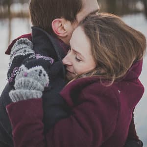 A woman in a burgundy coat hugging a man in a dark coat with snow in the background 