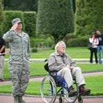 A soldier in green uniform stands saluting the flag next to a disabled vetern in wheelchair