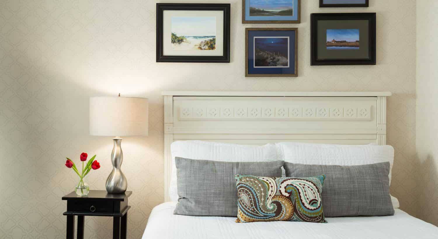 Bed with antiqued headboard, white bedding, gray pillows, light brocade wallpaper, and pictures hanging on the wall