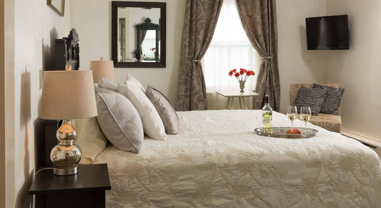 Bedroom with dark wooden headboard, cream bedding, silver tray with fruit and wine on the bed, upholstered chair, and flat-screen TV hanging on the wall