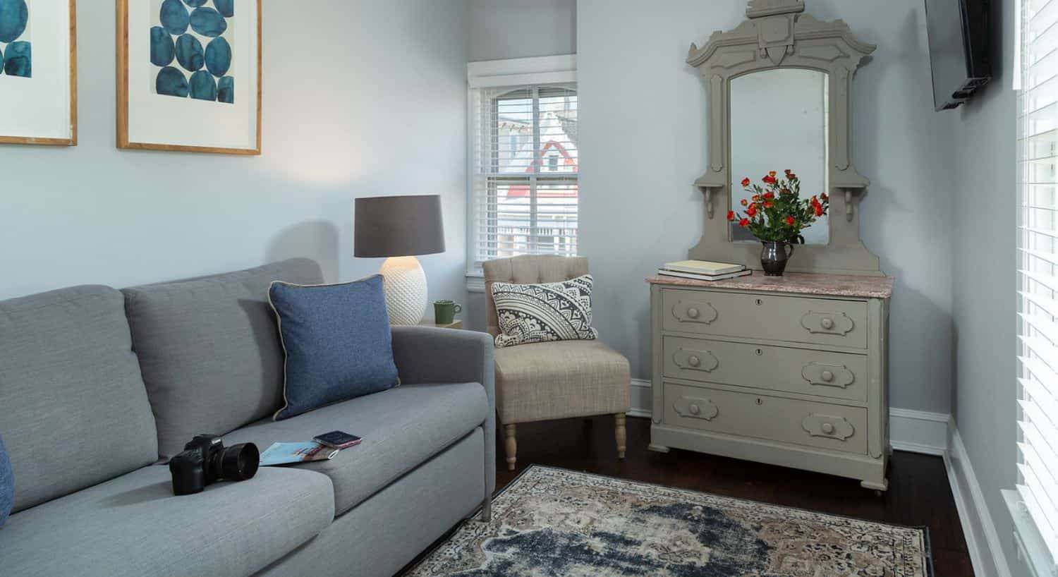 Sitting area with gray upholstered couch, digital SLR camera, denim pillow, hardwood flooring, light gray walls, and gray-painted dresser with mirror on top