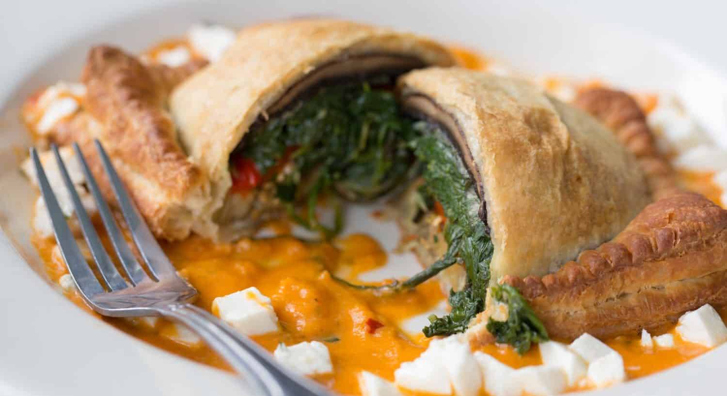 Close up view of a puffed pastry dish stuffed with spinach and vegetables surrounded by an orange cream sauce and mozzarella crumbles