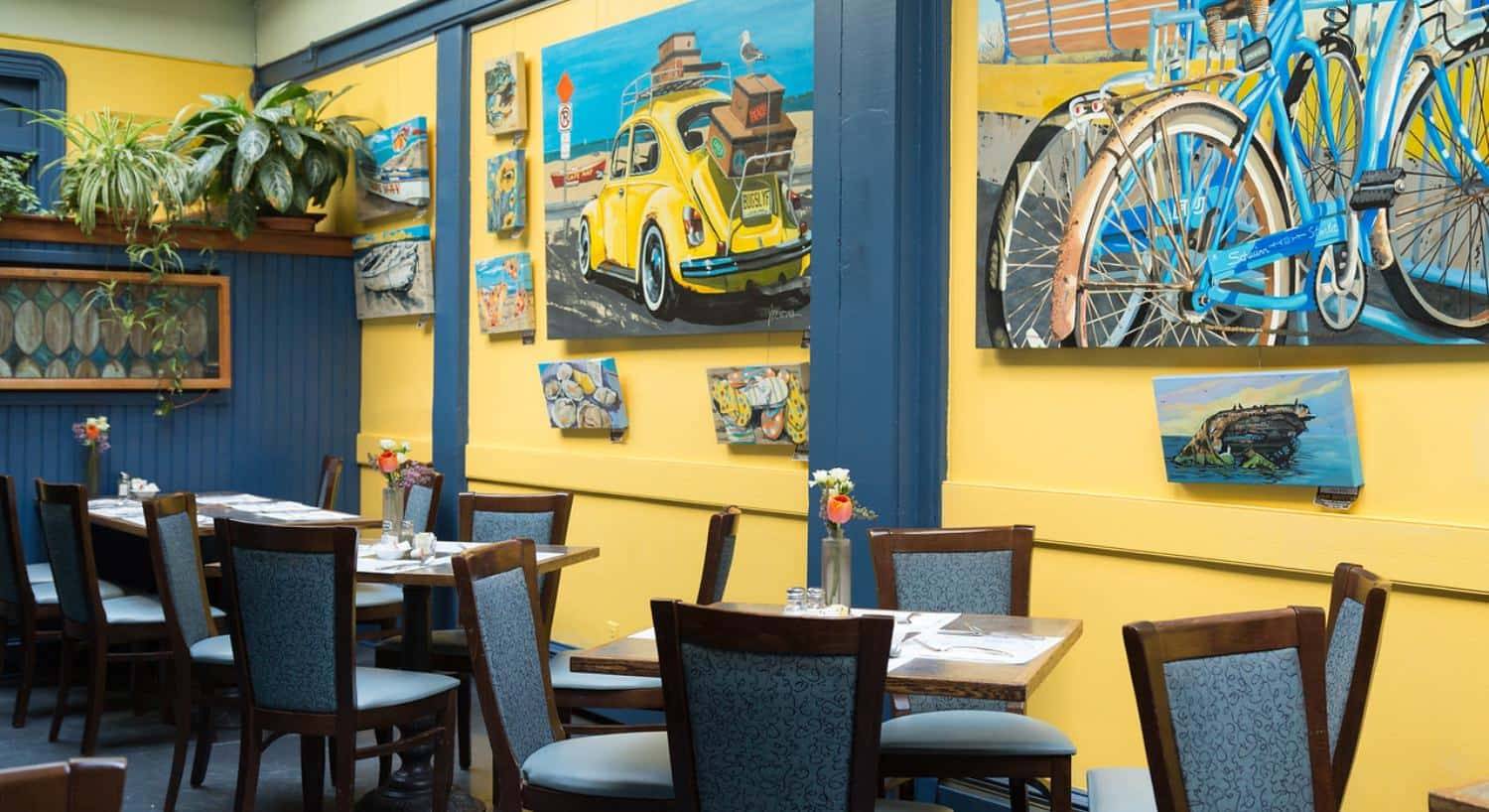 Dining area with dark wooden tables and chairs, yellow walls with blue trim, and large paintings on the walls