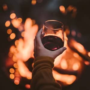 Person holding a glass of red wine while sitting in front of a fire at night