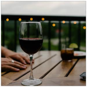 Glass of red wine on a wood table with patio lights in background. Image by serge-esteve-7517 www.unsplash.com